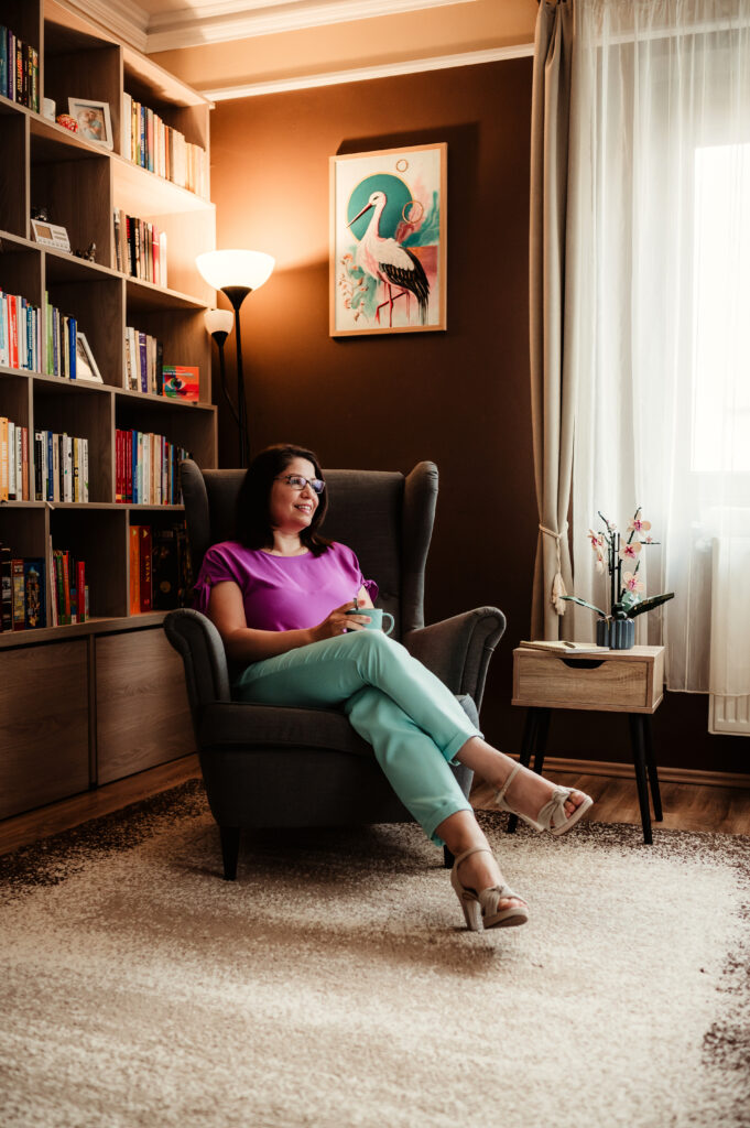 Kovács Marcsi mentor mosolyogva ül egy fotelben a könyvespolca előtt, kezében egy kávésbögrével.